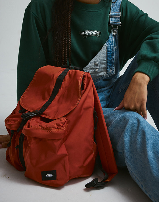 A young woman in a green Salton Pearls Crew Sweatshirt sits next to an orange backpack.