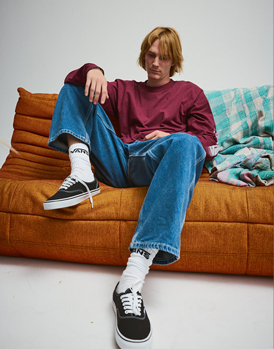 A young man sits on an orange sofa wearing a red Blur Drop Long Sleeve T-Shirt and black Authentic.
