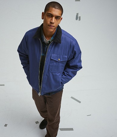 A young man poses in a photography studio wearing a deep twilight blue Skate Garage Reversible Jacket and chinos.
