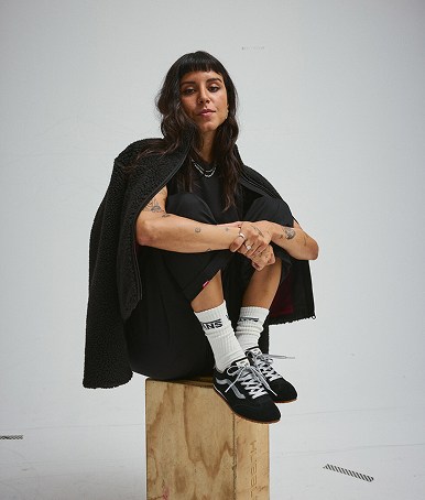 A young woman sits on a wooden box wearing Vans clothes and black and Super Lowpro.