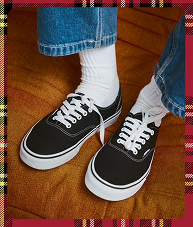 Gros plan sur une personne portant des Authentic noires avec un jean bleu et des chaussettes montantes Vans blanches, assise sur un sofa orange.
