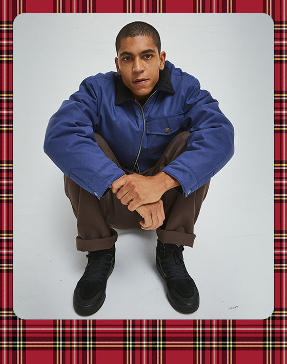 A photograph surrounded by a plaid holiday border of a young man wearing a deep twilight blue Skate Garage Reversible Jacket and chinos.