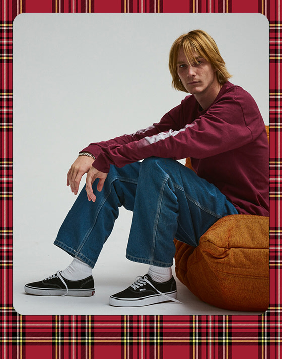 A photograph surrounded by a plaid holiday border of a young man wearing black Authentic and a red Blur Drop Long Sleeve T-Shirt.on a skateboard cross-legged inside a warehouse. They are wearing a black and white Vans trucker hat, a short sleeve green plaid shirt with a white thermal underneath, brown jeans, and Old Skool sneakers in black.