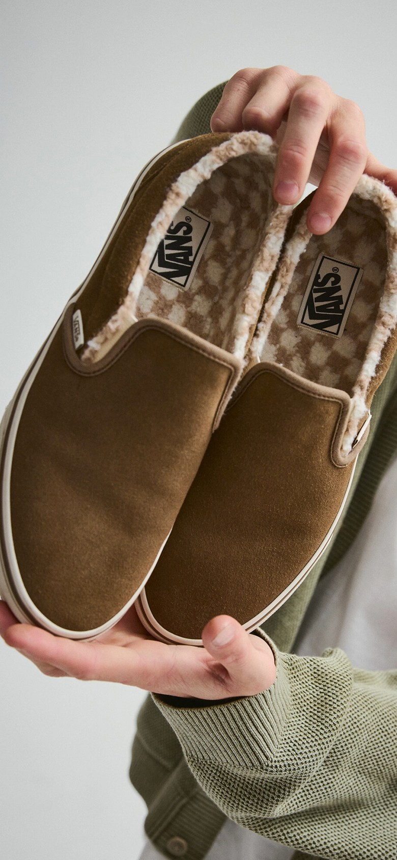 A young man holds up a pair of brown Classic Slip-On with fuzzy Checkerboard lining.