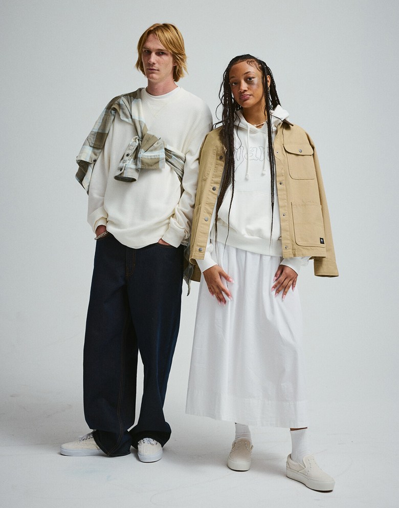 A young man and woman pose in a photography studio wearing new Vans clothes, including the Drill Chore Jacket and the Larkspur Flannel Shirt.