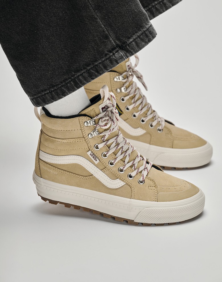 Close-up of a man sitting on a box wearing tan Sk8-Hi Insulated boots with black jeans in a photography studio.