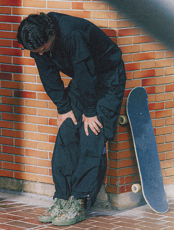 A man leaning against a brick wall, wearing the OTW by Vans X OAMX x WTAPS camo sk8 hi shoes and all black track suit with a skateboard standing up next to him.