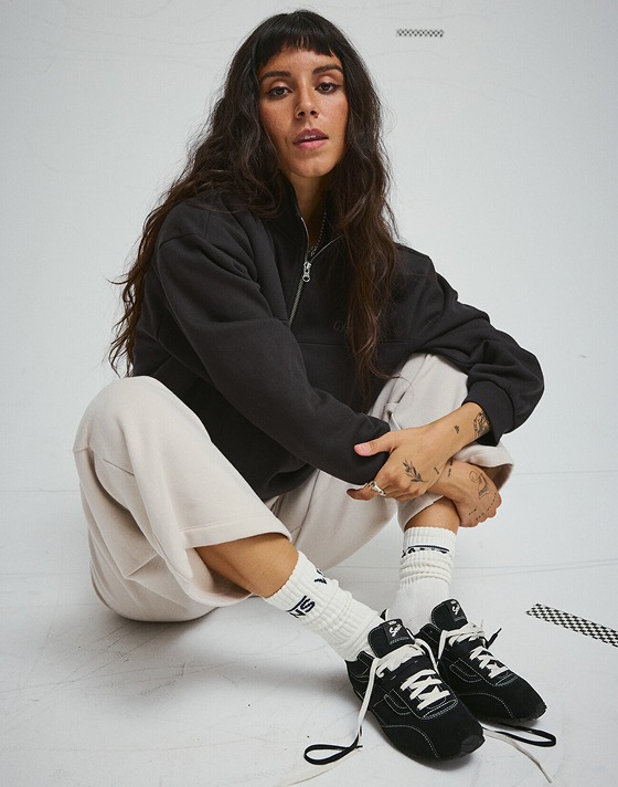 A young woman sits on the floor of a photography studio wearing black Premium Super Lowpro Trainer.