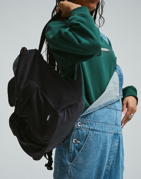 A holds a black Vans backpack wearing blue denim Sirelle overalls in a photography studio.
