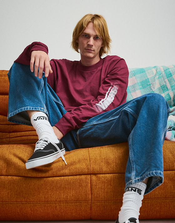 A young man poses on an orange sofa wearing black and white Authentic, blue jeans, and a red Blur Drop Long Sleeve T-Shirt.