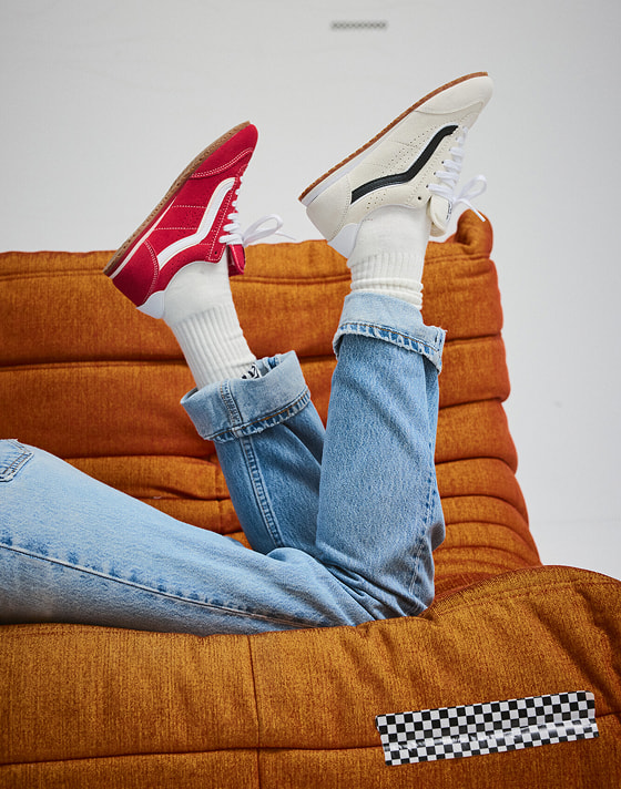 Someone lying on an orange sofa wearing a marshmallow white Super Lowpro on one foot and a racing red Super Lowpro on the other with blue jeans.