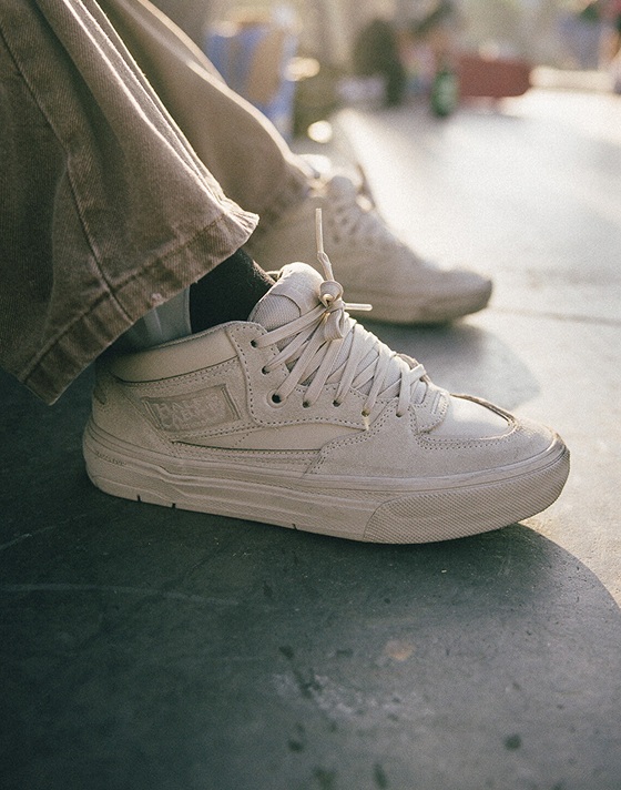 Close-up of someone sitting and wearing white Half Cab shoes with gray jeans at a skate park.