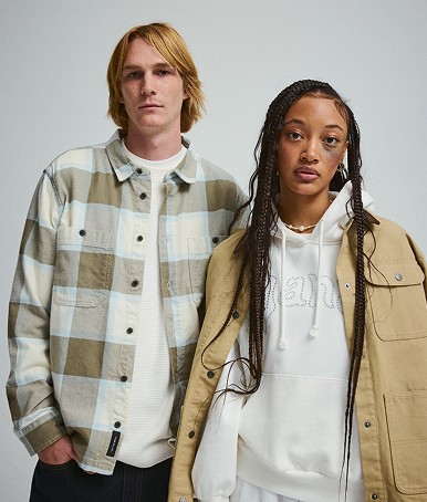 A young man in a Larkspur Plaid Flannel Shirt stands next to a young woman in a tan Drill Chore Coat in a photography studio.