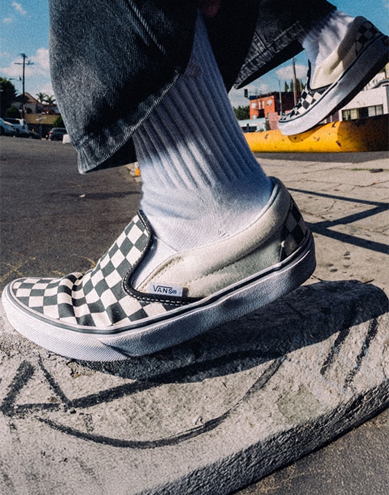Close-up of the black and white Checkerboard Classic Slip-On in a parking lot styled with black jeans and white crew socks.