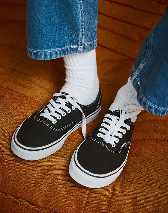 Someone wearing black and white Authentic on an orange sofa with blue jeans and white crew socks.