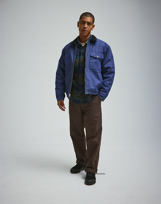 A young man wearing a deep twilight blue Skate Garage Reversible Jacket and chinos in a photography studio.