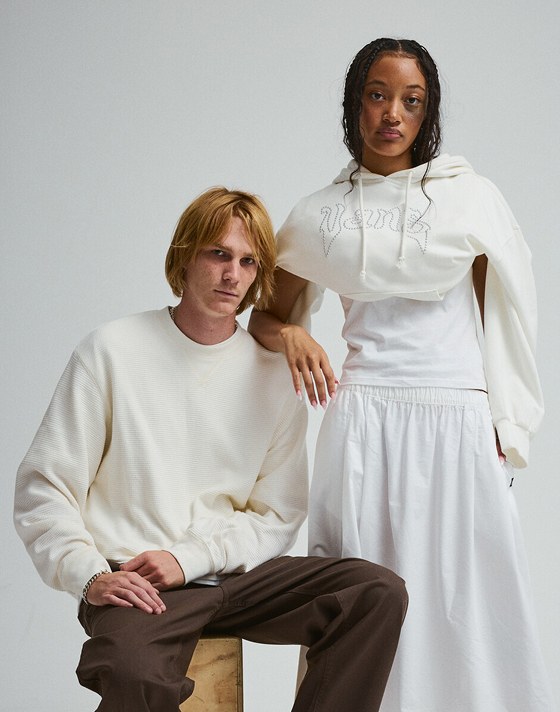 A young man and woman pose in a photography studio wearing a white crewneck and white hoodie against a white wall.
