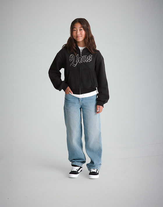 A young girl poses in a photography studio wearing a black Kids Shining Zip Hoodie and Kids Sirelle Denim Puddle Pants.