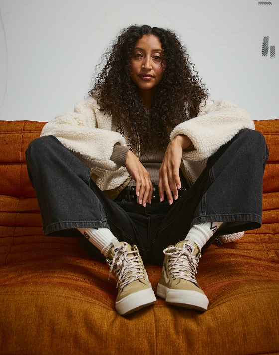 A young woman sits on an orange sofa in a photography studio wearing a pair of tan Sk8-Hi Waterproof Insulated boots.