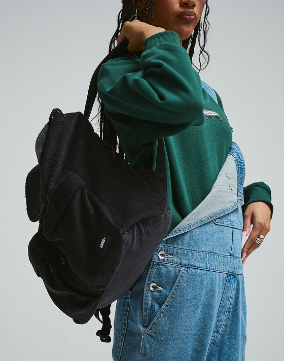A young woman wearing the Emerald Crew Salton Pearls and Sirelle Overalls putting a black backpack over her shoulder in a photography studio.