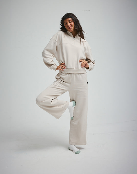 A young woman poses in a photography studio wearing a taupe mist gray Script Logo Mock Neck Sweatshirt and Drop V Sweatpants.