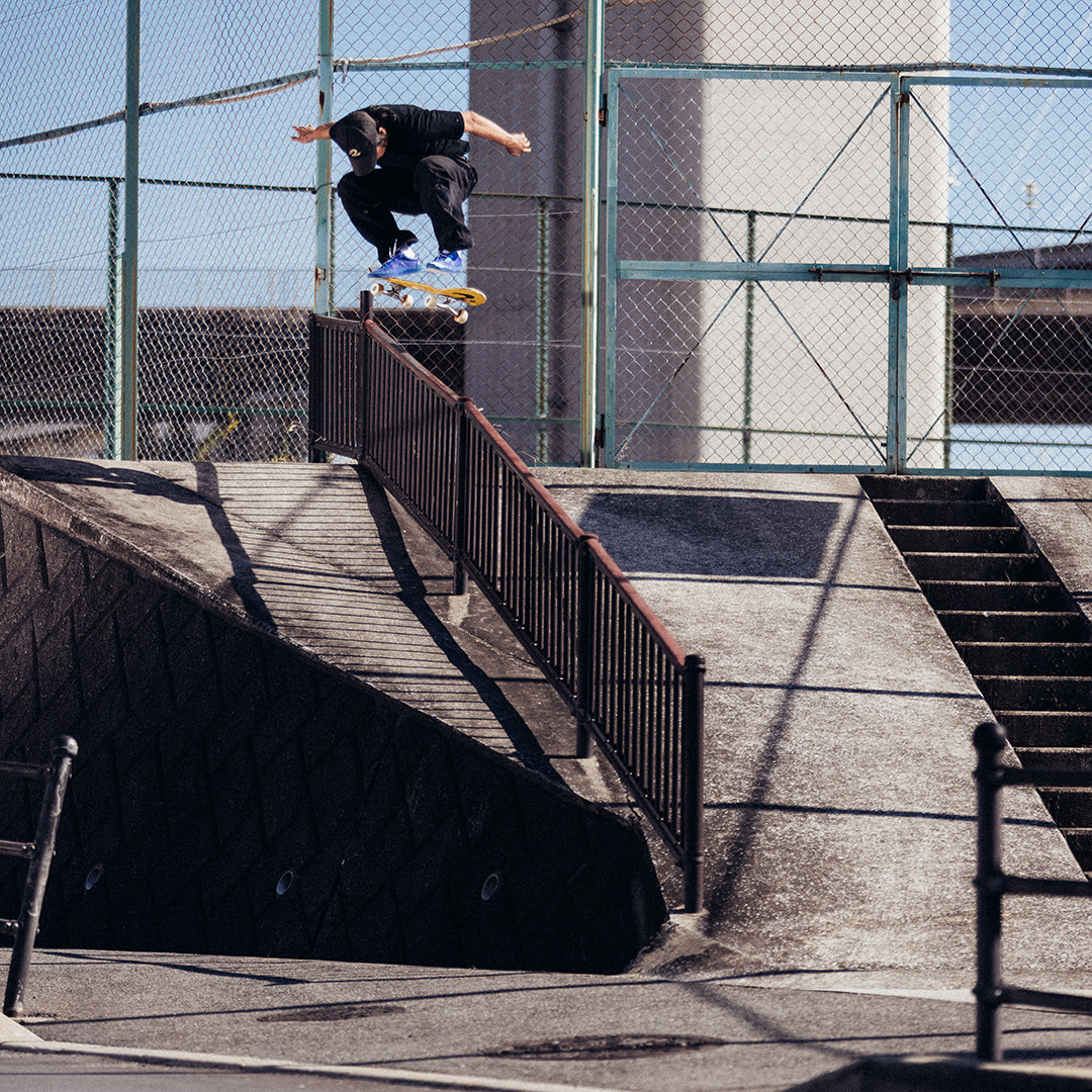 Skateboarder performing a high ollie over a handrail at an urban spot, captured mid-air against a chain-link fence backdrop.