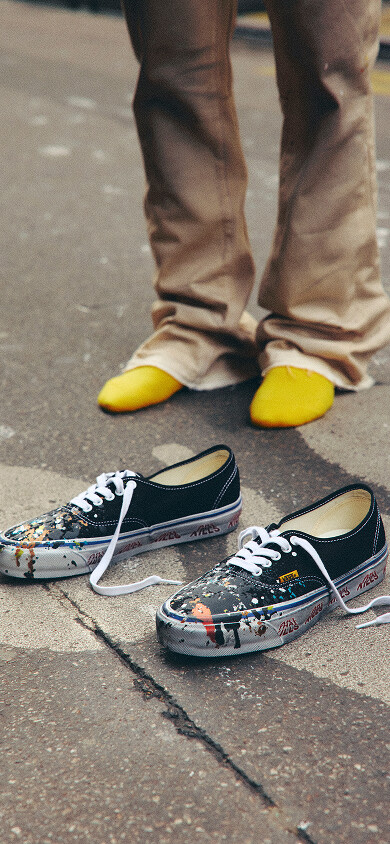 A person standing in socks next to OTW by Vans X Gallery Department Authentic 44 with Paint Splatter Canvas Black.