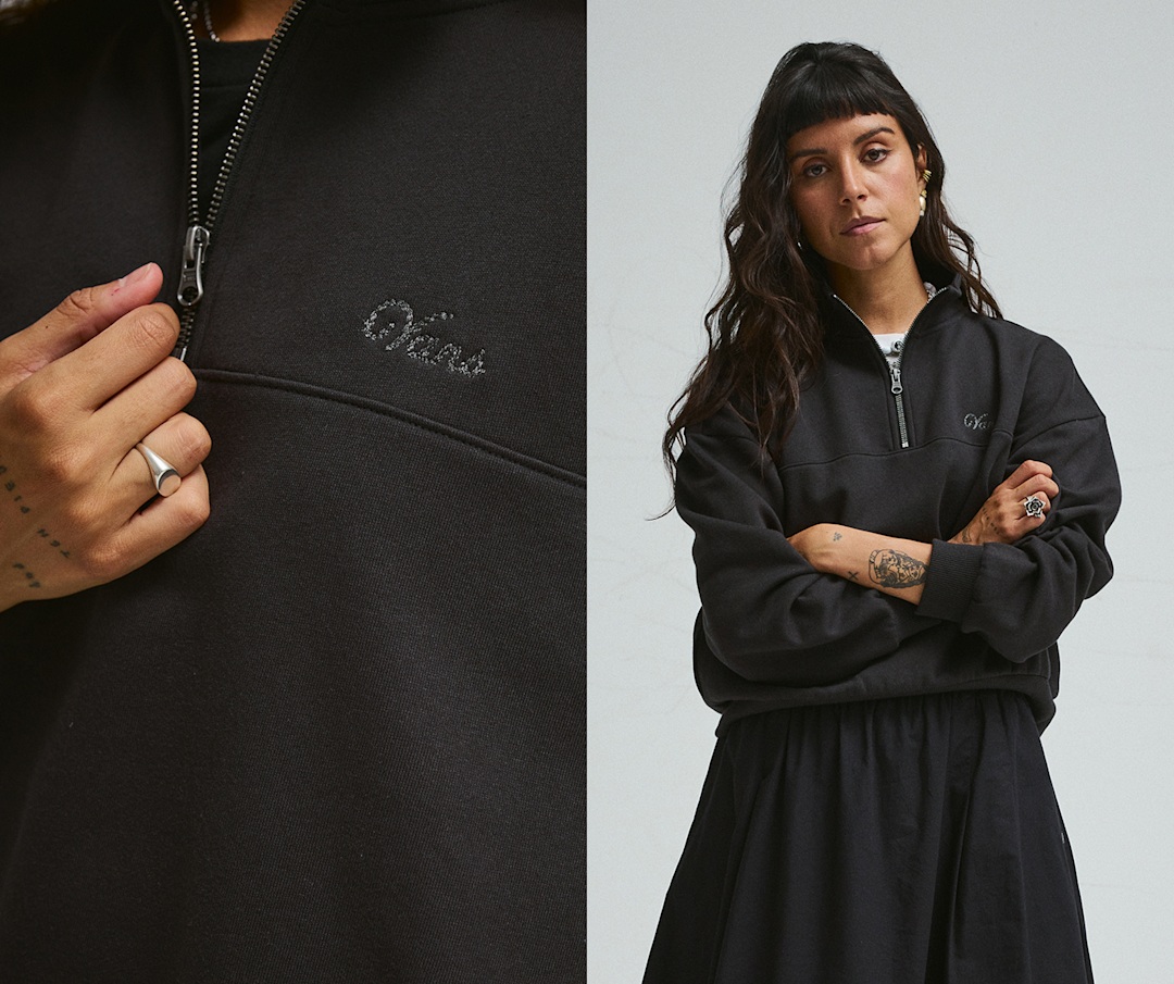Une jeune femme croise les bras en portant un chandail à col montant noir avec le logo Script, dans un studio photo.