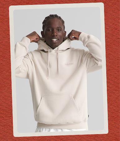 A young man wearing a taupe mist gray Vans 66 Pullover Sweatshirt on a red holiday background.