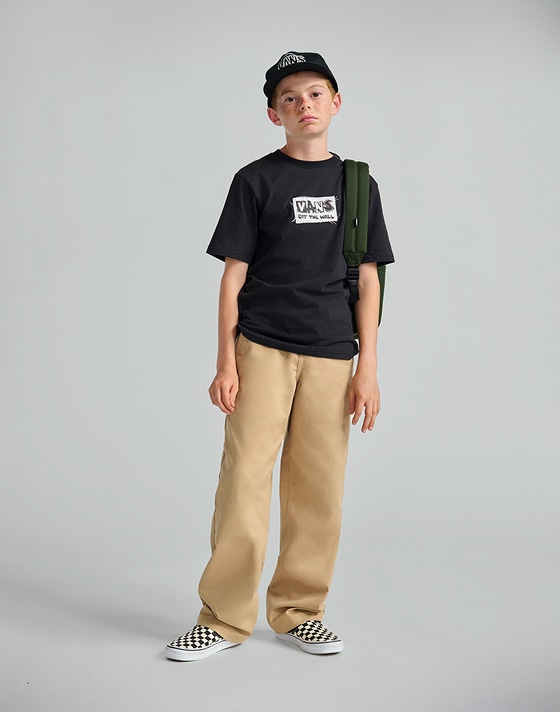 A young boy poses in a photography studio wearing a black Kids DIY FX T-Shirt, a pair of tan kids’ chinos, and Checkerboard Classic Slip-On.