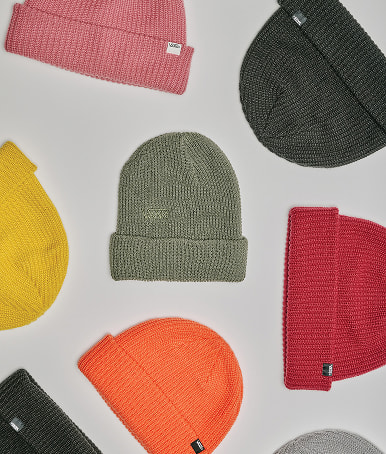 Multiple beanies are laying on the floor of a grey photography studio, each one is a different color such as orange, red, green, yellow, pink, and grey.