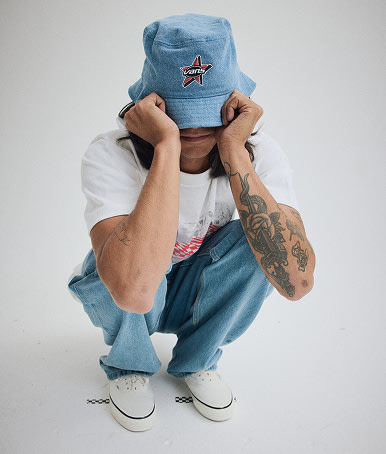 Person squatting in a grey photography studio wearing a Vans Denim Bucket Hat.