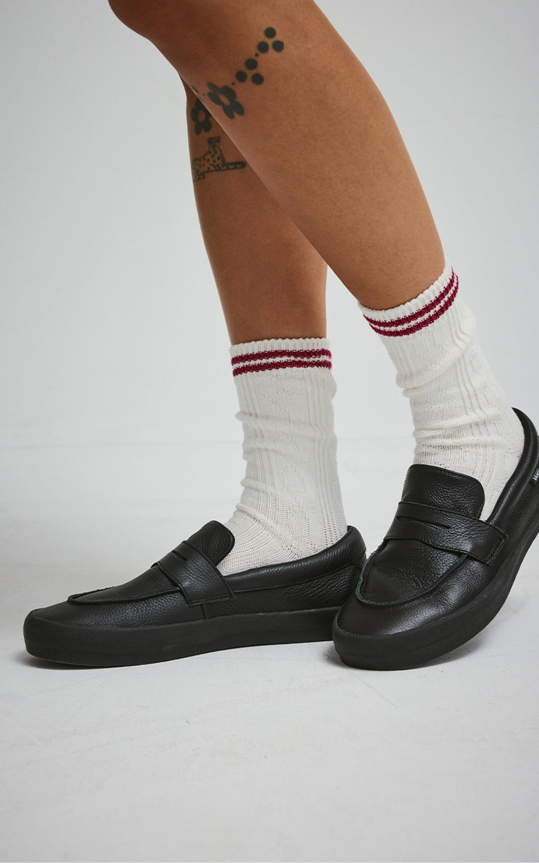 Close up from knee down. Someone is wearing The Skate Loafer in all-black with a pair of white crew socks with two red stripes at the top.