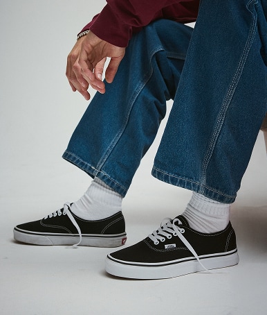 A waist-down shot of a young man sitting on an orange sofa wearing black and white Authentic shoes with blue jeans and white Vans crew socks.