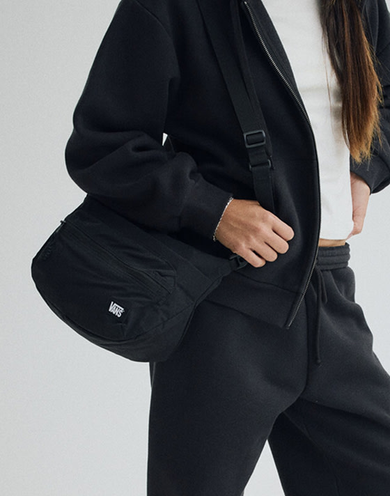 Image of a person wearing an all black zip up with a white shirt underneath and black sweats. They are carrying a black bag on their shoulder.
