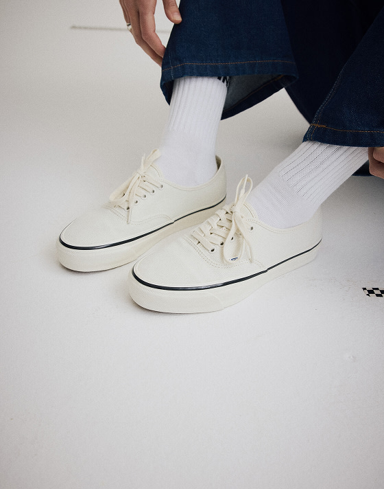 Close up of someone wearing the Premium Authentic Shoe in Marshmallow White while sitting on the ground of a grey photography studio.