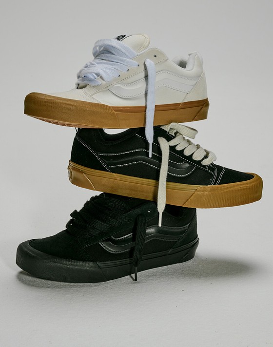 A Blanc De Blanc White Knu Skool shoe, a black gum Knu Skool shoe, and a black Knu Skool shoe stacked on top of each other in a photography studio.