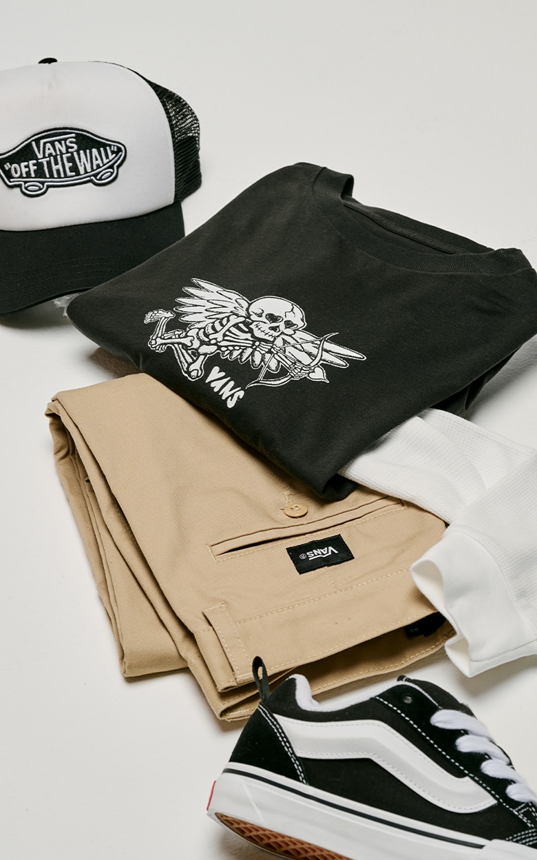 A pair of kids’ black and white Knu Skool, Kids Authentic Chino Loose Pants, a black tee, and a hat on the floor of a photography studio.