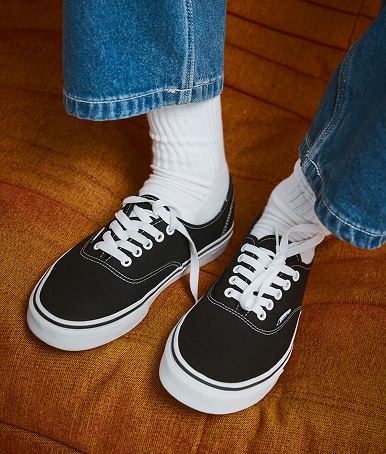 Someone wearing black and white Authentic on an orange sofa with blue jeans and white crew socks.