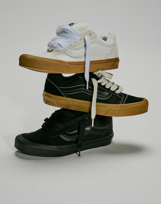 A Blanc De Blanc White Knu Skool shoe, a black gum Knu Skool shoe, and a black Knu Skool shoe stacked on top of each other in a photography studio.