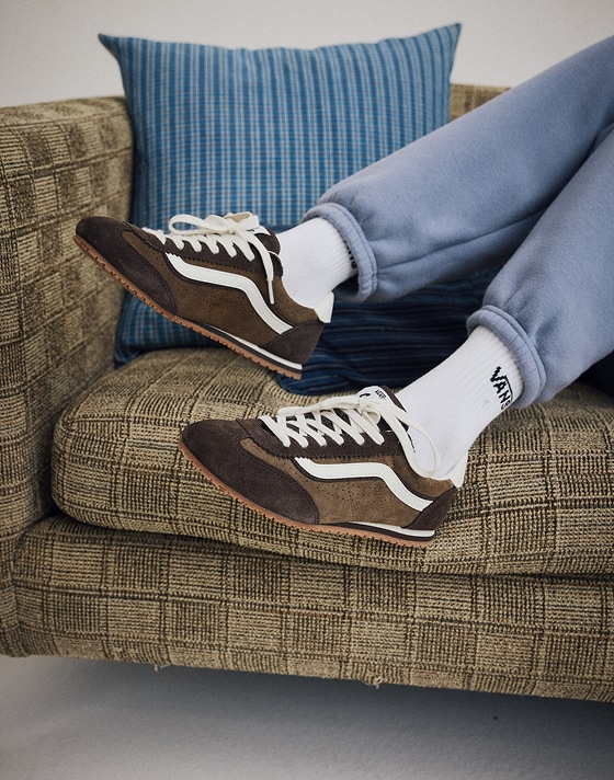 A knee-down shot of someone wearing demitasse brown Super Lowpro, light blue sweats, and white Vans crew socks on a retro-looking sofa.