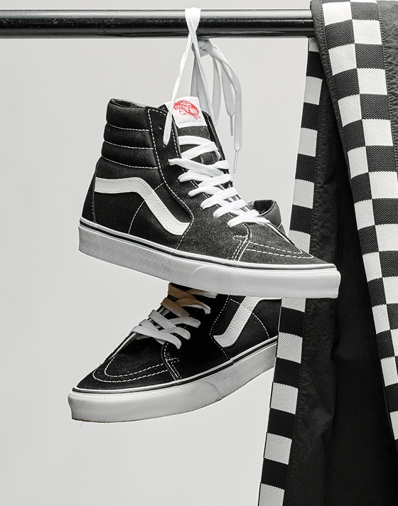 A pair of black and white Sk8-Hi hang by their laces from a metal clothing rack in a photography studio.