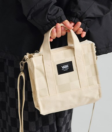 A woman holding an oatmeal beige Lil Pergs Checkerboard Tote Bag behind her back wearing all black in a photography studio.