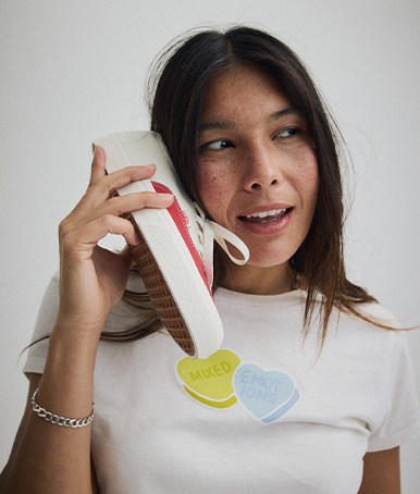 A young woman holds up a red and white Authentic to her face like a phone wearing a Mixed Emotions t-shirt from the Vans Valentine’s Day Collection.