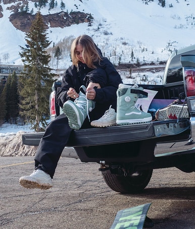 A young woman laces up her olive gray Women's Luna Pro Snowboard Boot in the bed of a pickup truck in the parking lot of a ski resort.