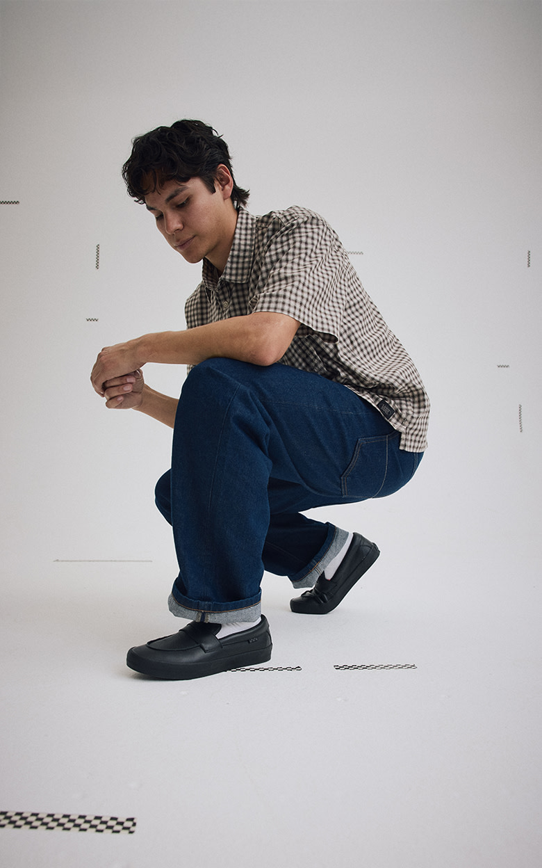 Medium shot of Nick Michel of the Vans Skate Team wearing the black leather Skate Loafer with jeans and a plaid button in a grey photography studio.
