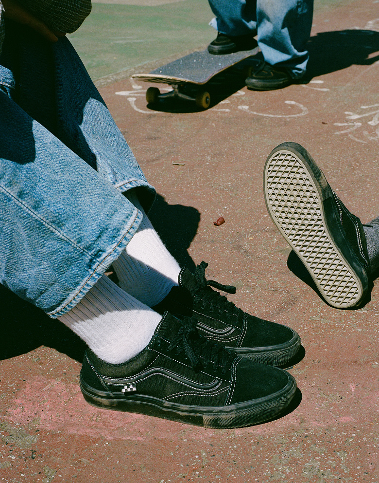 Medium knee down shot of three people wearing the all black Skate Old Skool shoe on the side of an old tennis court.
