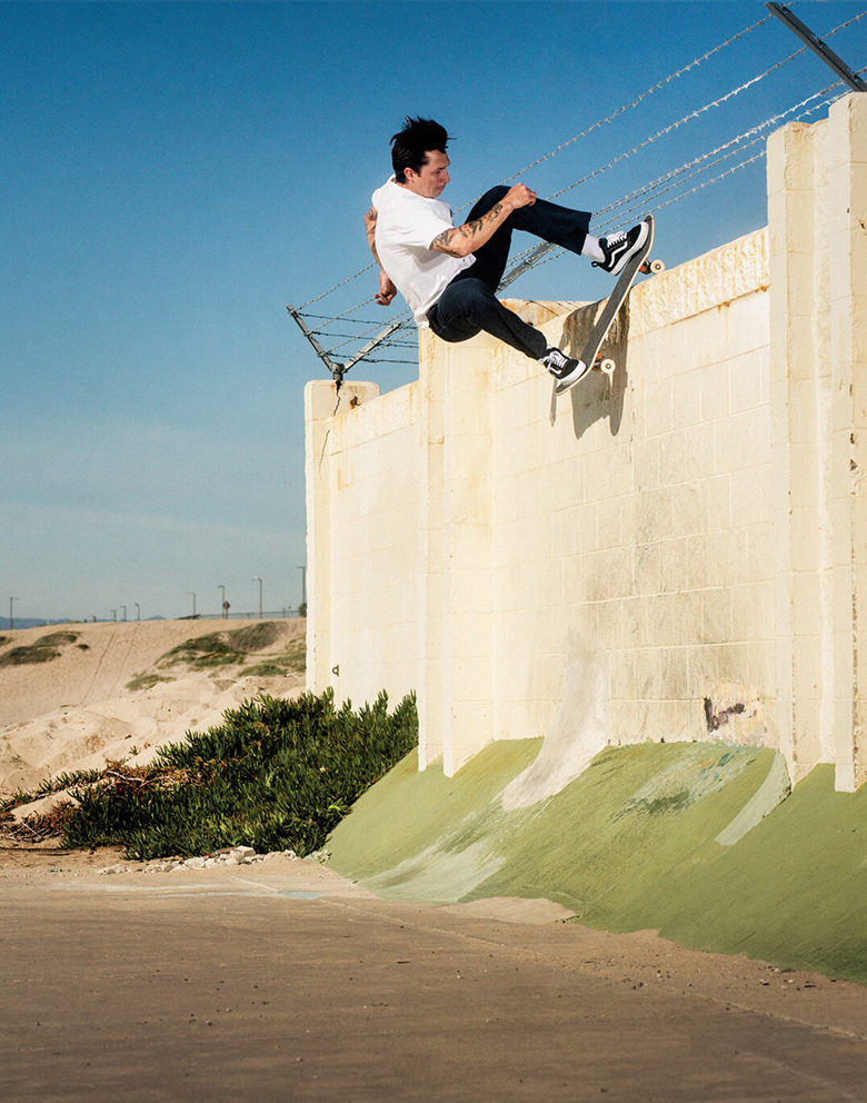 Vans Skateboarder Elijah Berle is skating on the edge of a cement wall with barbed wire fencing. The background is blue sky and desert.