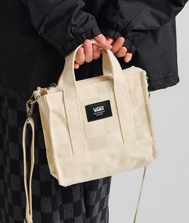 A woman holding an oatmeal beige Lil Pergs Checkerboard Tote Bag behind her back wearing all black in a photography studio.