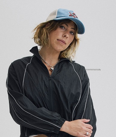 Vans Surf Team Rider Karina Rozunko poses in a photography studio wearing a Steve Navy NYLA Track Jacket and a Marked Jockey Trucker hat.